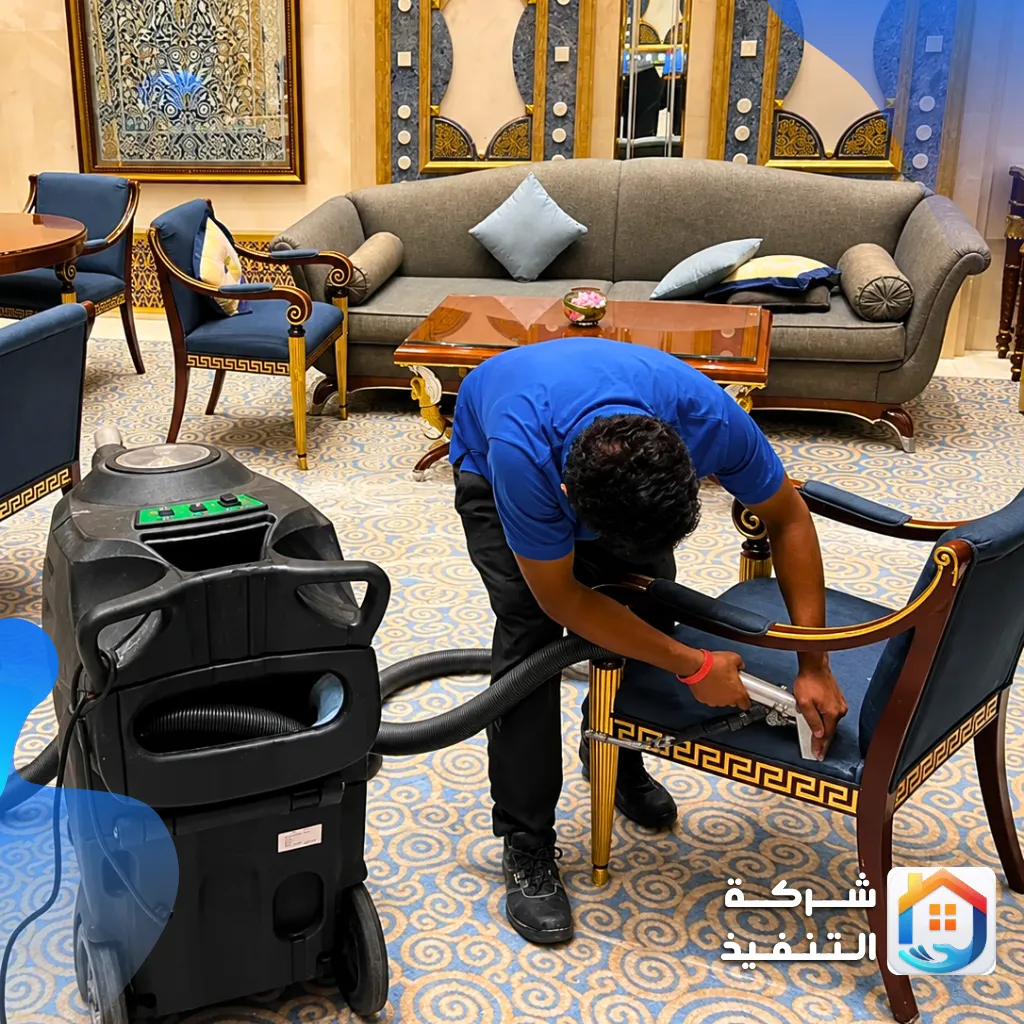 فني ينظف كرسي انتريه كحلي بإطار خشبي مذهب بماكينة شفط في صالة استقبال، أثناء تنظيف كنبات الرياض